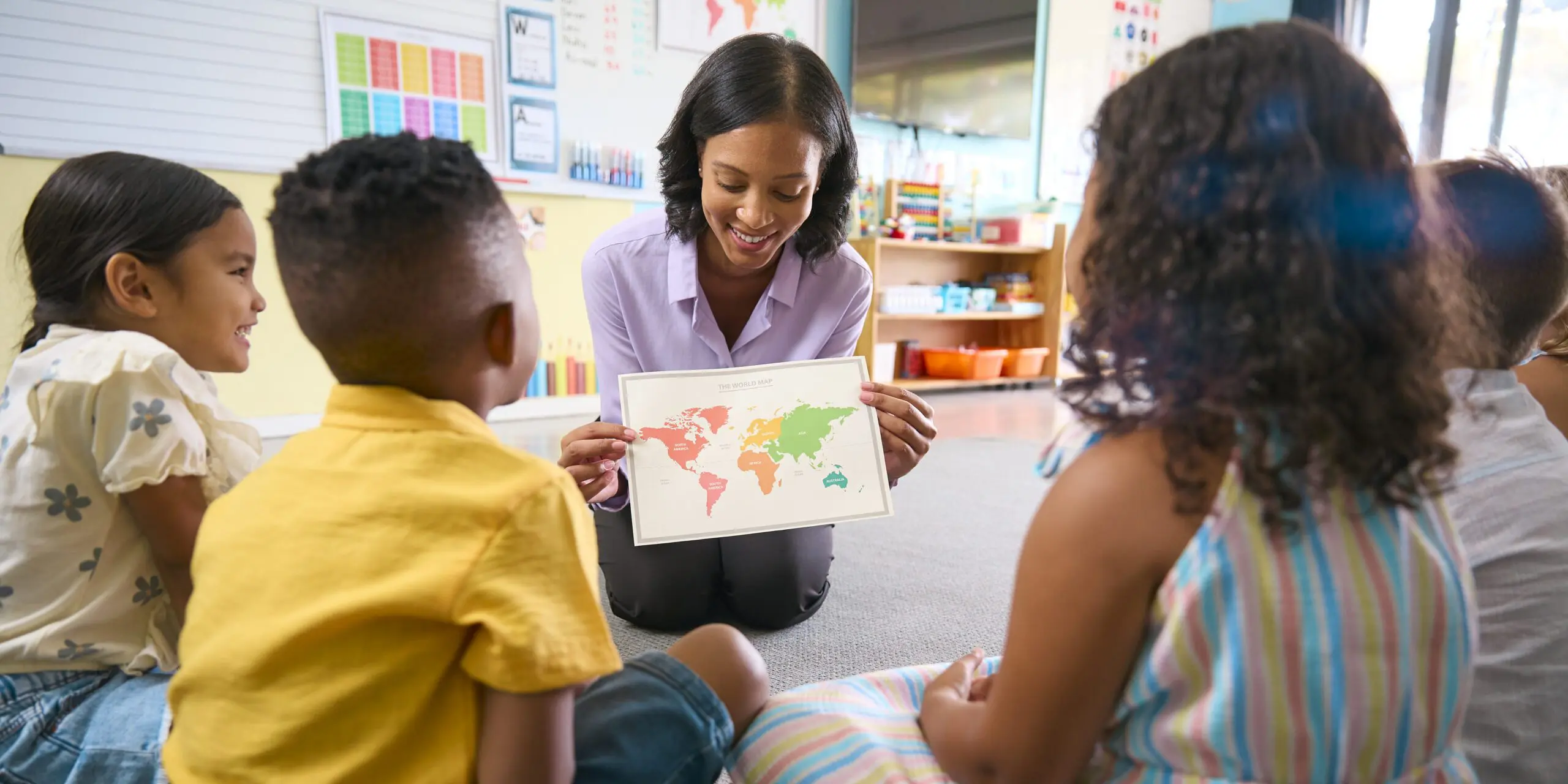 Female Primary Or Elementary School Teacher In Multi-Cultural Class With Map In Geography Lesson